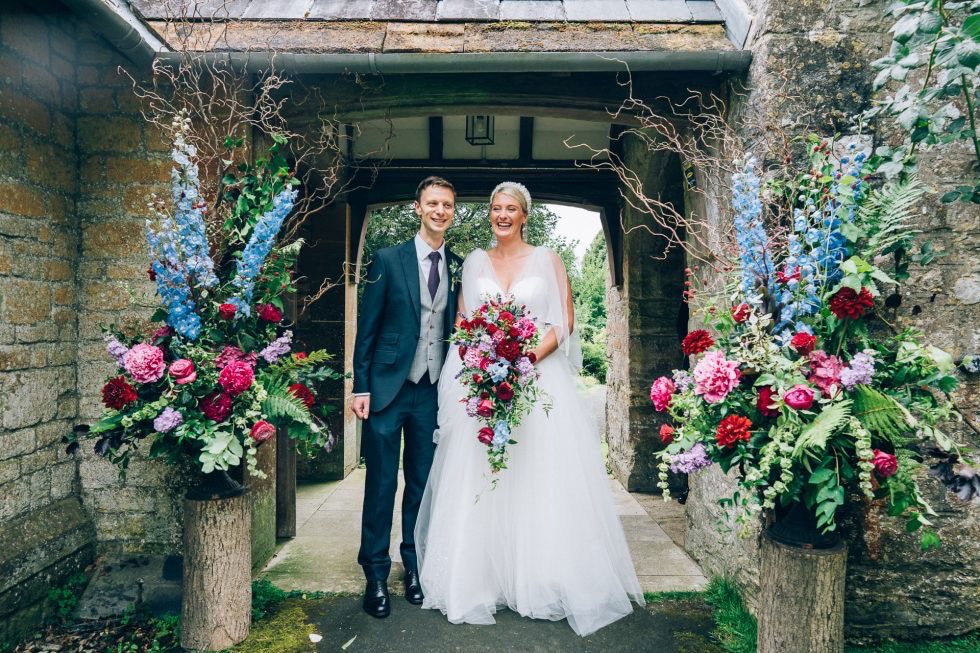 Bride and groom in a stone doorway flanked by two tall arrangements of delphiniums, roses and foxgloves