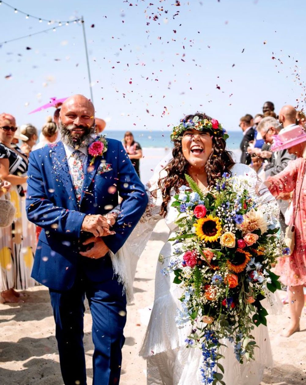 Bride and groom showered with confetti on a beach, the bride carrying a trailing sunflower bouquet and wearing a flower crown