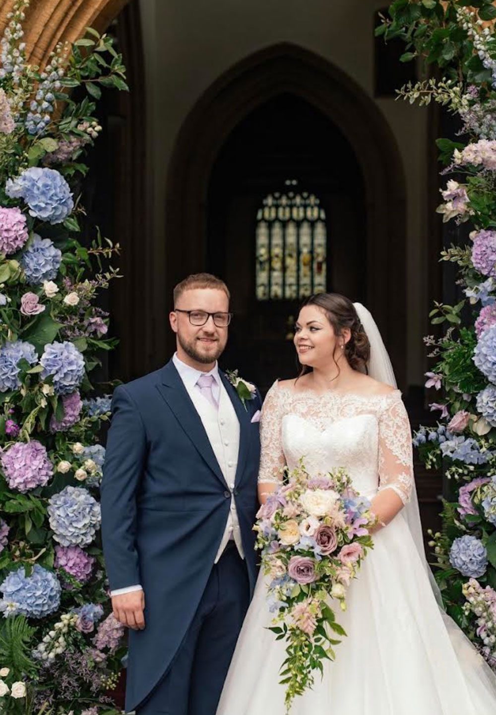 Bride with a trailing bouquet and groom standing in a doorway arch of hydrangeas
