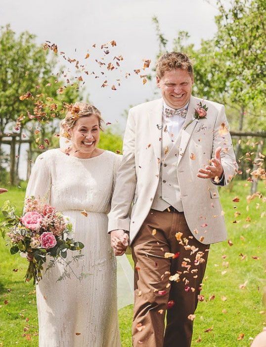 Bride and groom walking through confetti in an orchard, the bride carrying a pink peony bouquet