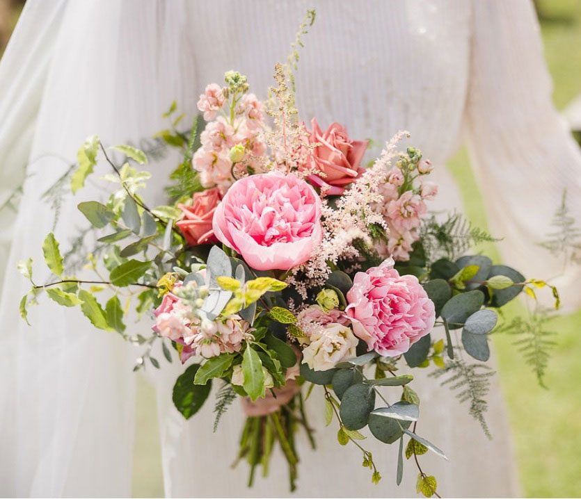 Hand-tied bouquet of pink peonies, roses and astilbe held against a wedding dress