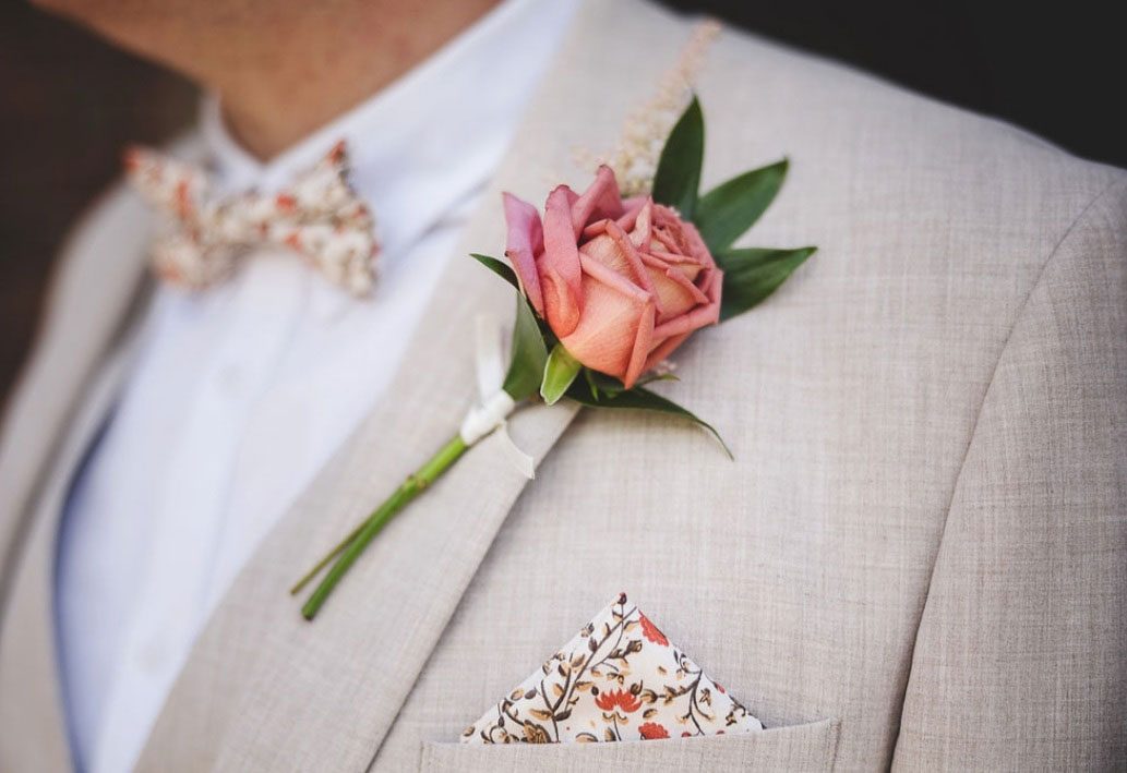 Coral rose buttonhole pinned to a pale linen jacket with a floral bow tie