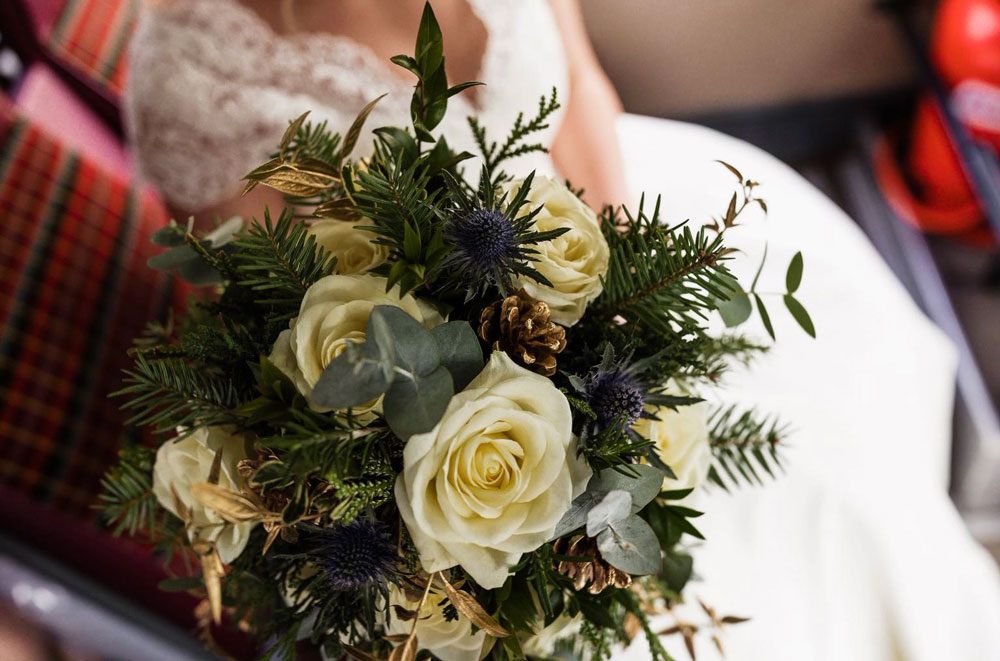 Winter bouquet of ivory roses, thistles and pine held against a tartan wrap